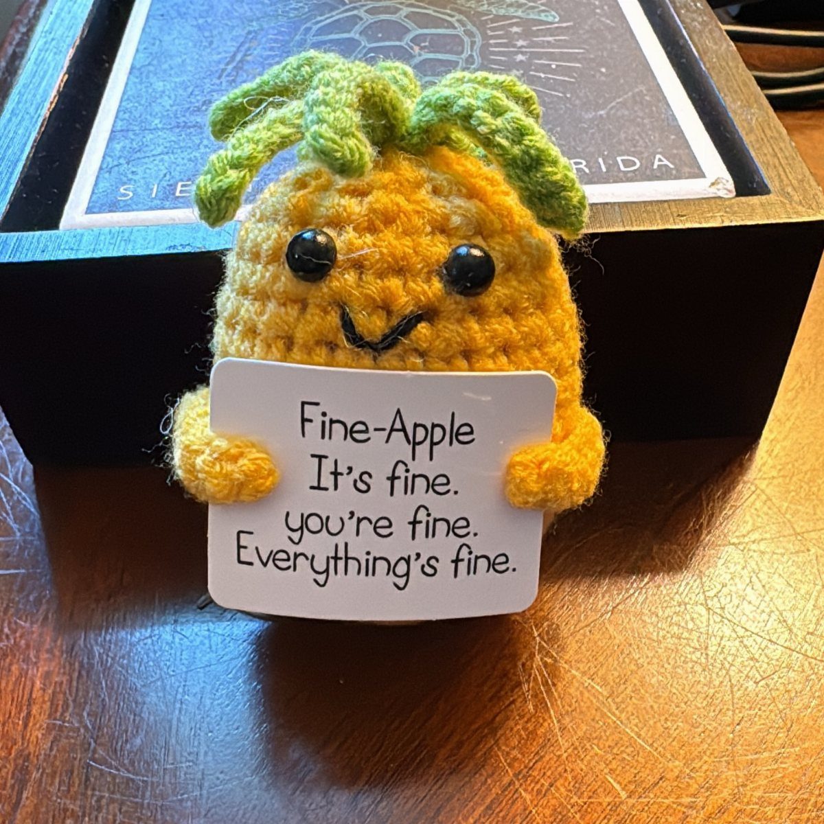 Crocheted pineapple plush holding a sign with a supportive message on wood.