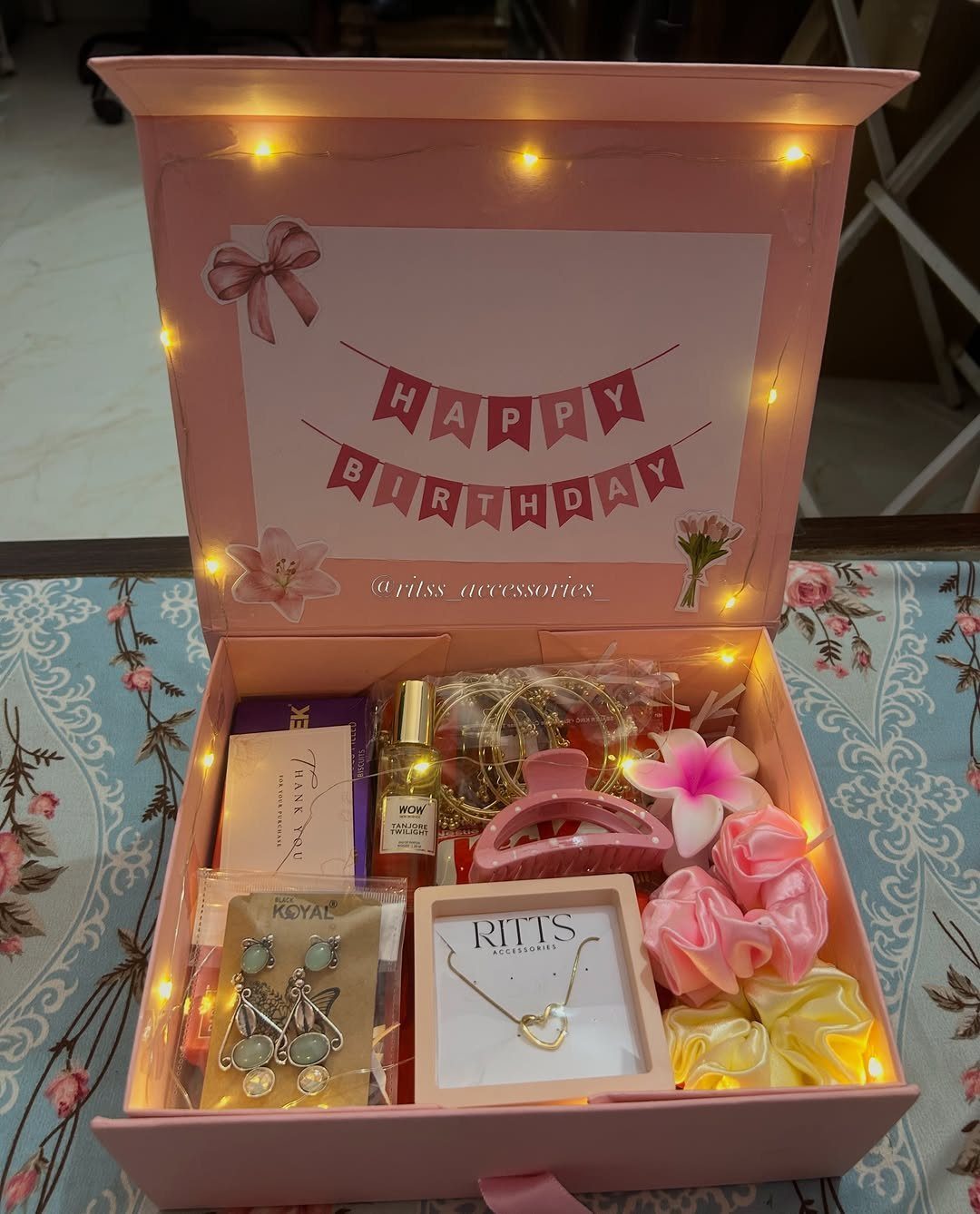 Pink birthday gift box filled with jewelry, perfume, and hair accessories with fairy lights.