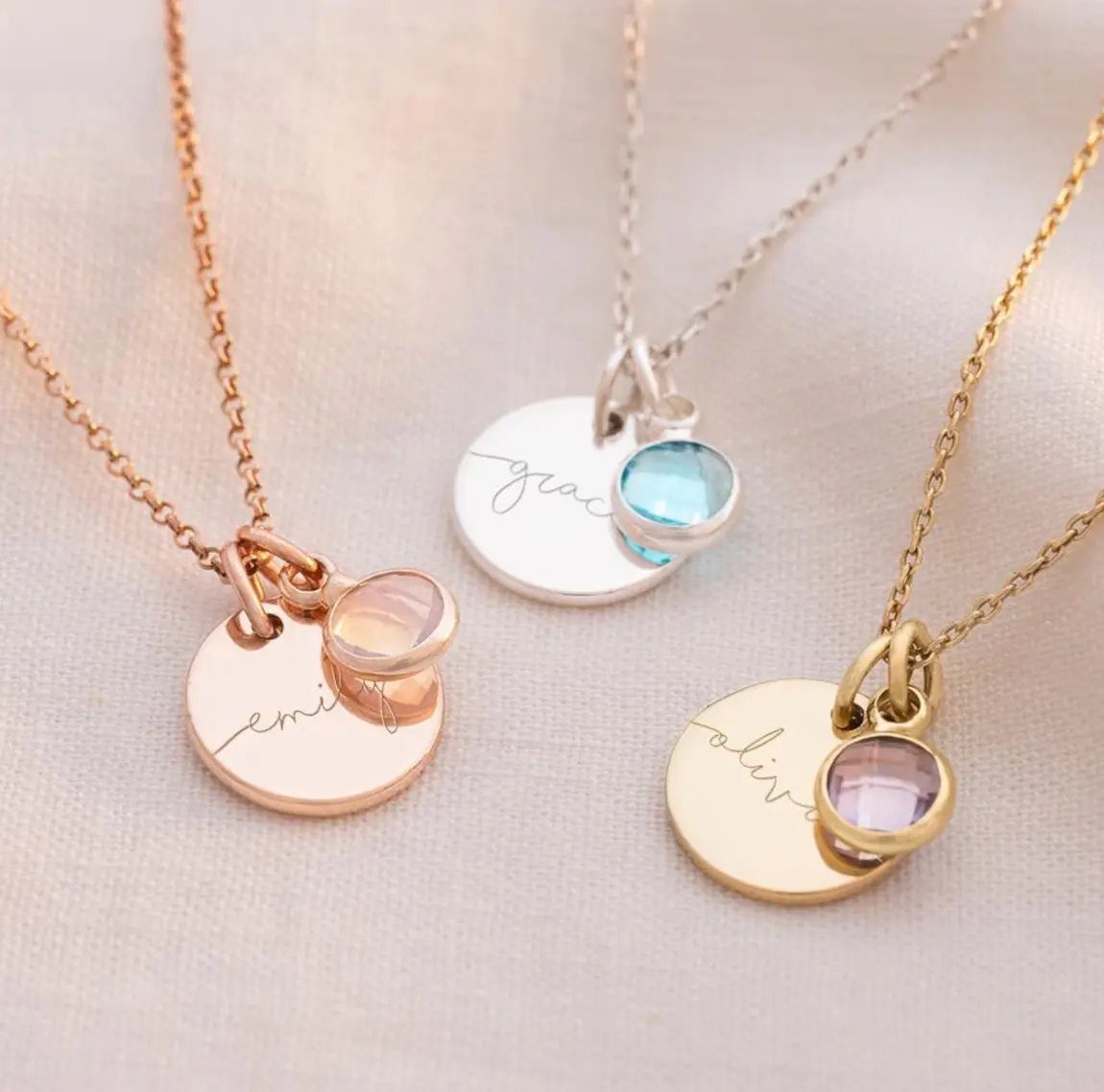Three personalized necklaces with name pendants and colorful birthstone gems on white fabric.