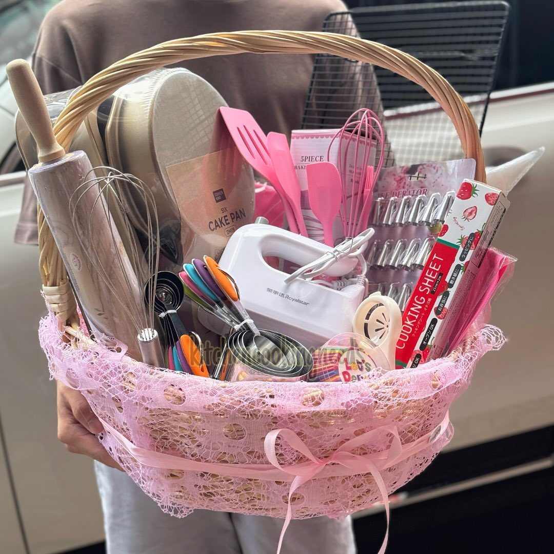 Pink gift basket filled with various kitchen tools and baking accessories held by person.
