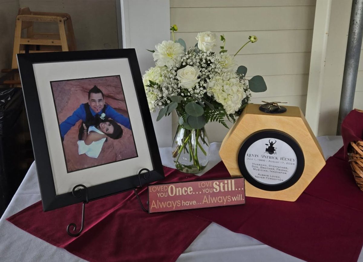 Memorial table with framed couple photo, white floral arrangement, and wooden urn.