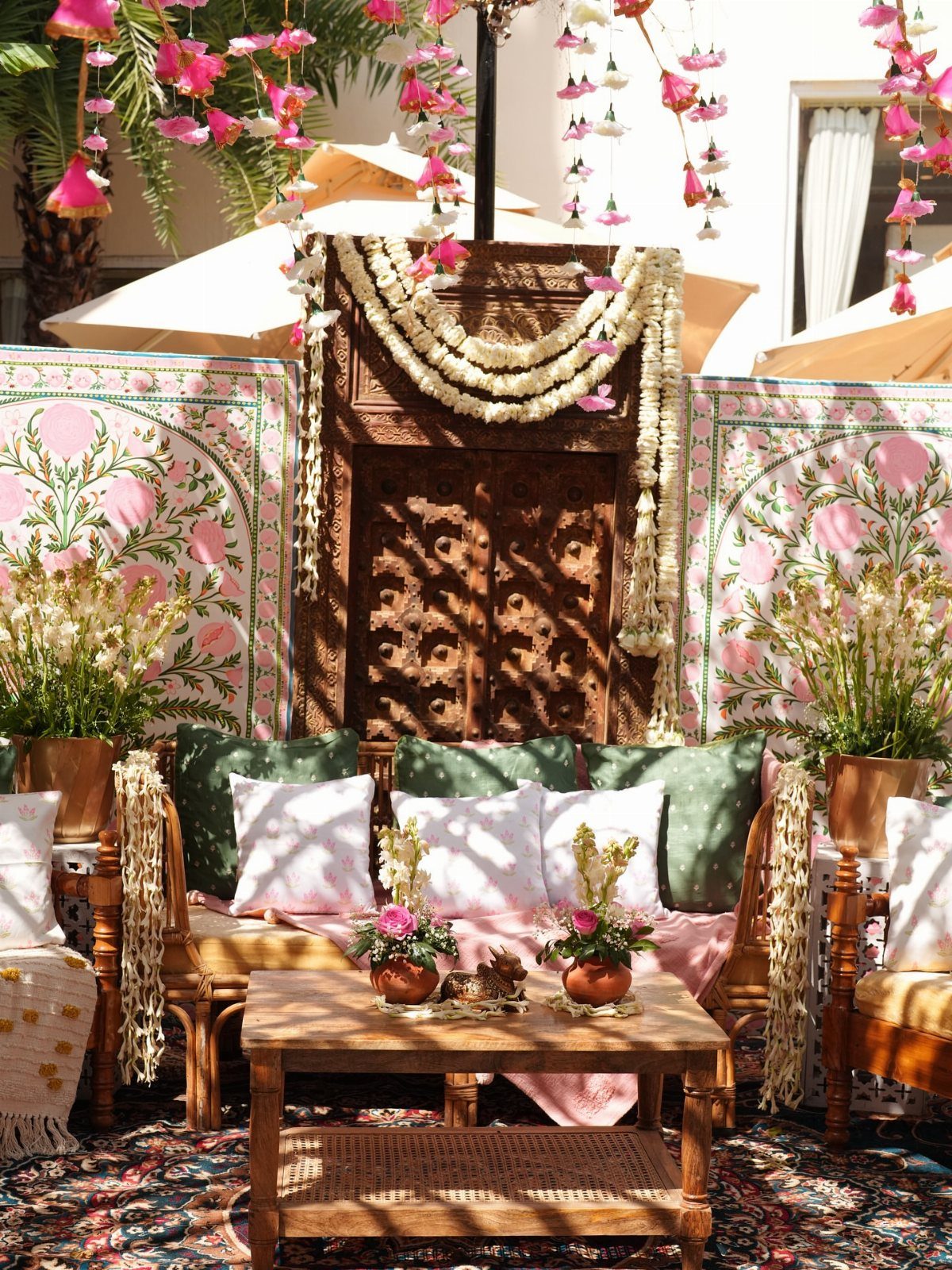 Outdoor lounge area with wooden furniture, colorful cushions, and floral decorations on a rug.