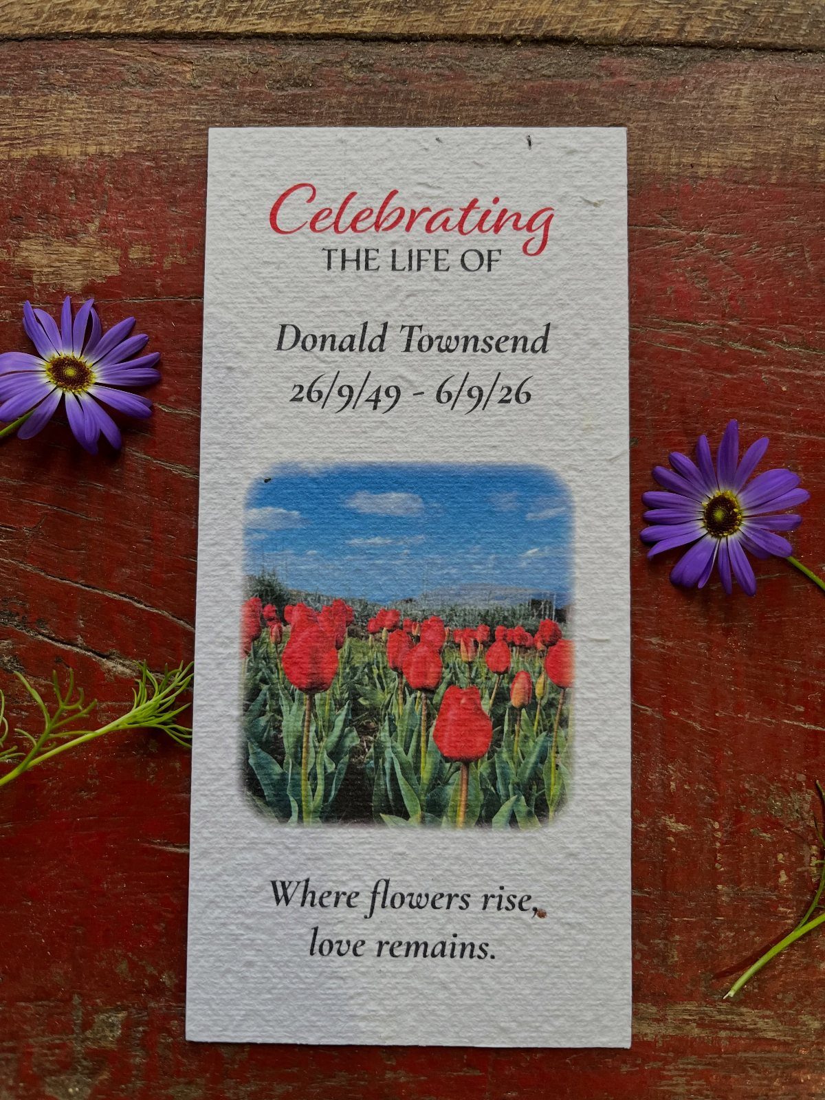 Memorial bookmark with poppy photo and text resting on red wood with purple flowers.