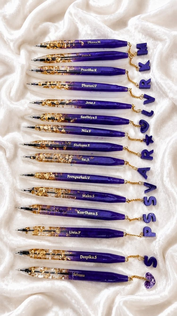 Purple resin pens with gold flakes and personalized letter charms on white fabric.