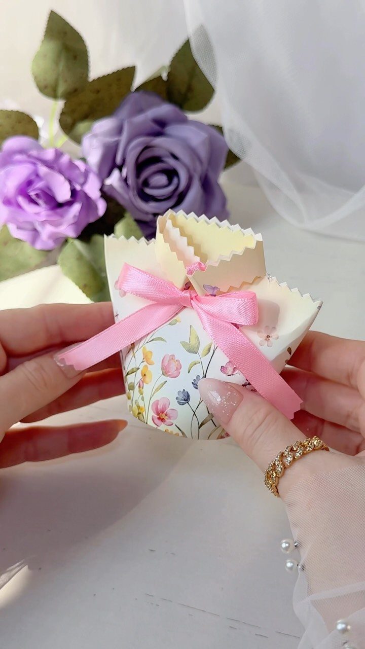 Floral gift box with pink ribbon held by hands near purple roses.