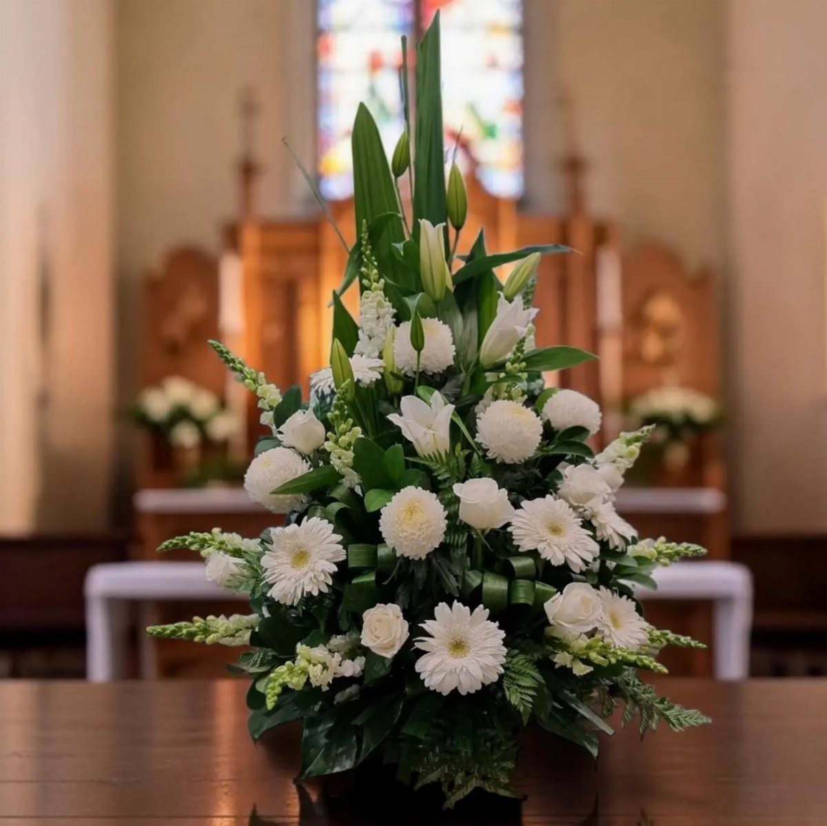White floral arrangement with lilies and daisies on a wooden church table.