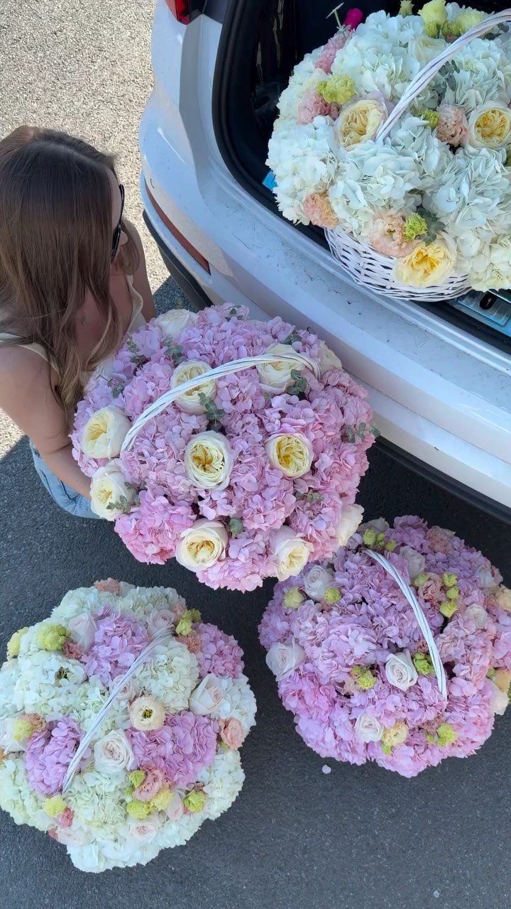Four large floral baskets with pink, white, and yellow flowers near a car.