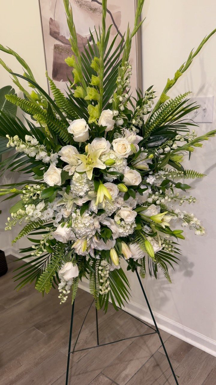 White floral standing spray with roses and lilies on a black metal easel.