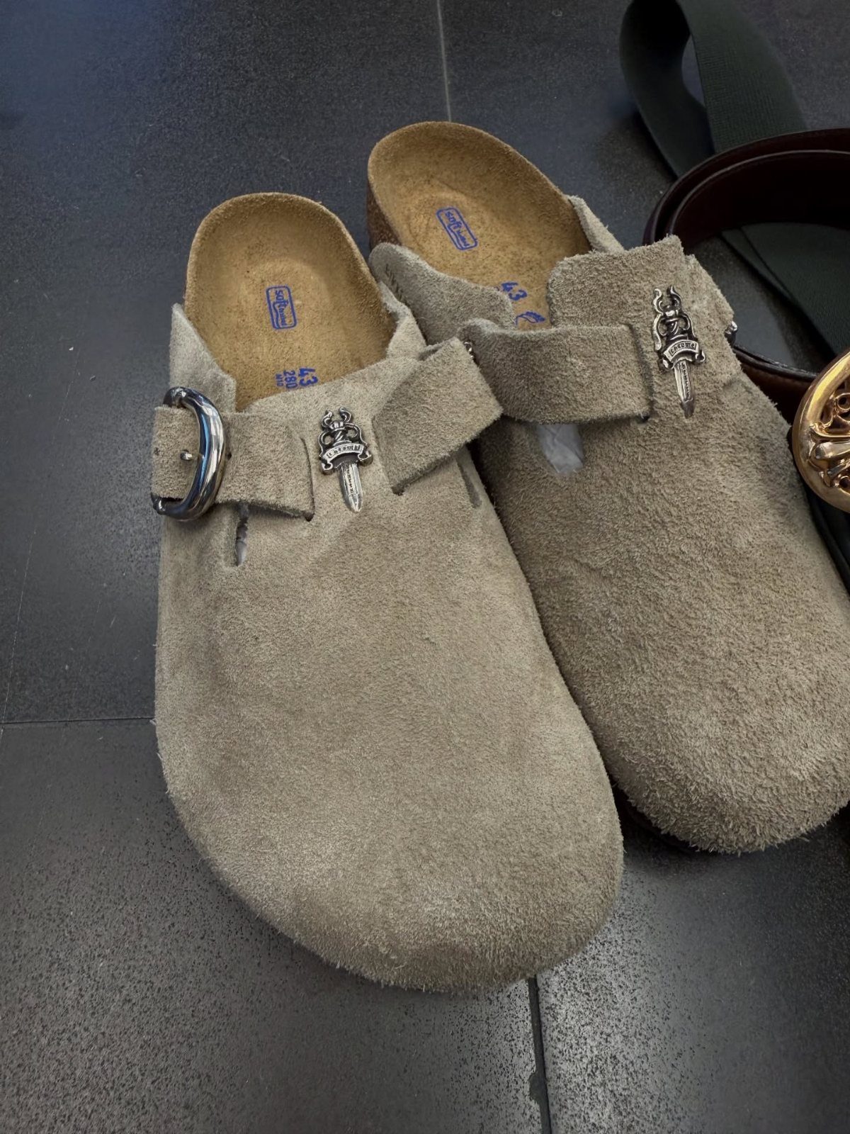 Beige suede clog shoes with silver buckles on a dark grey tiled floor