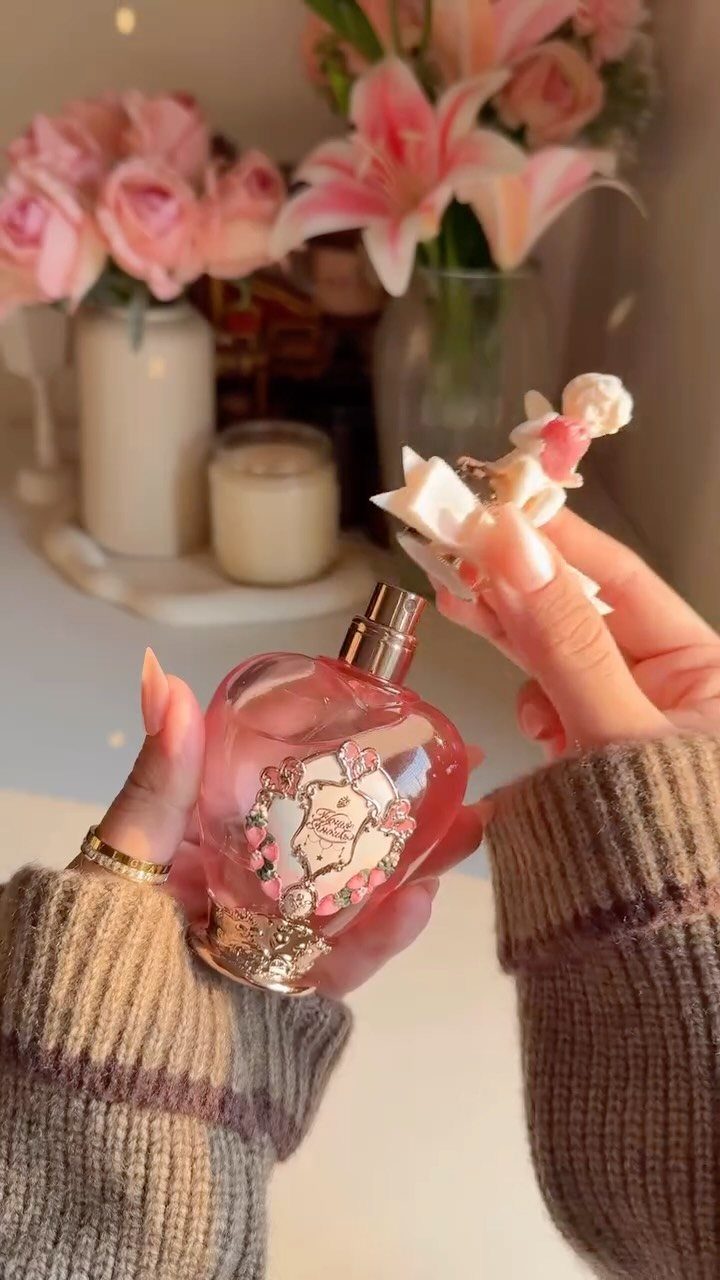 Pink heart-shaped perfume bottle held by hands against a background of pink flowers.