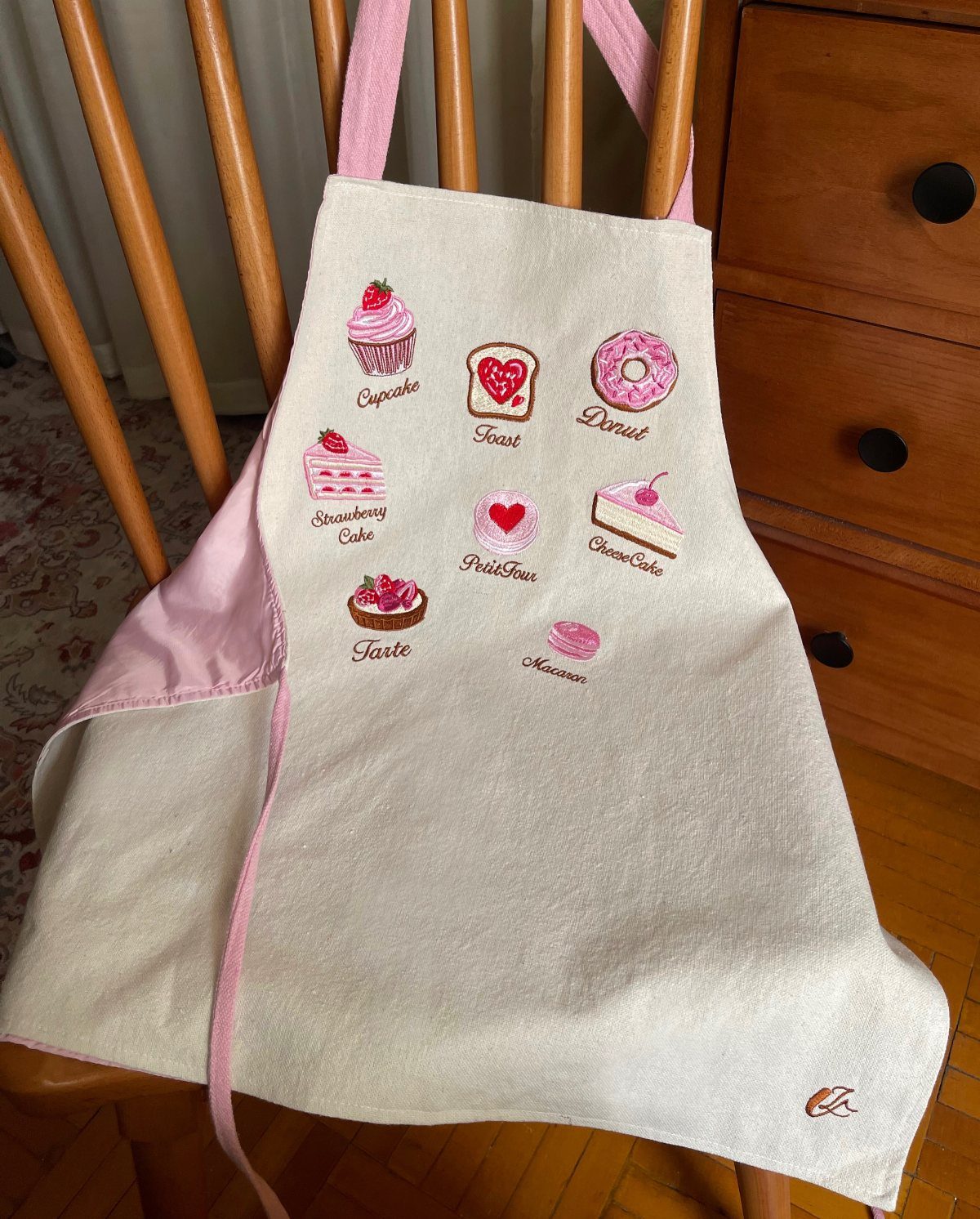 Cream apron with pink straps and embroidered dessert icons hanging on a wooden chair