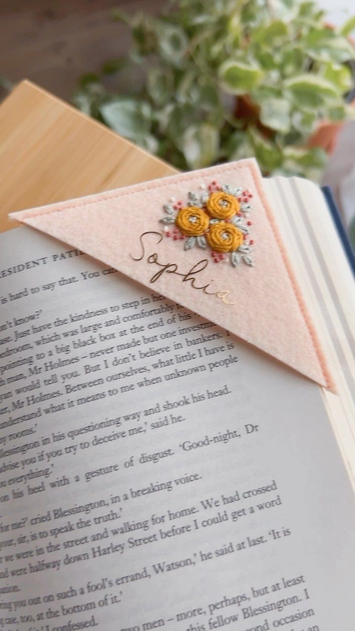 Pink fabric corner bookmark with yellow embroidered flowers and name on a book page