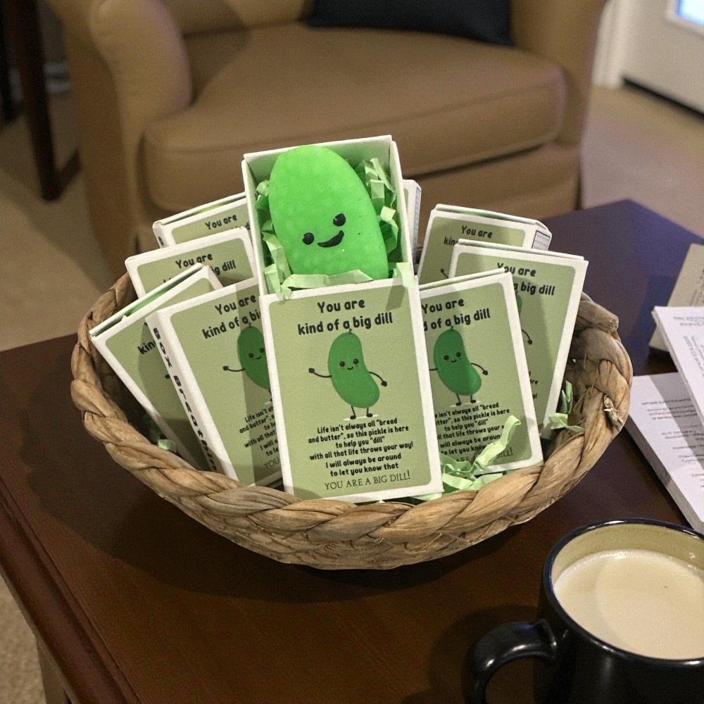Small green plush pickles in gift boxes inside a woven basket on a table.