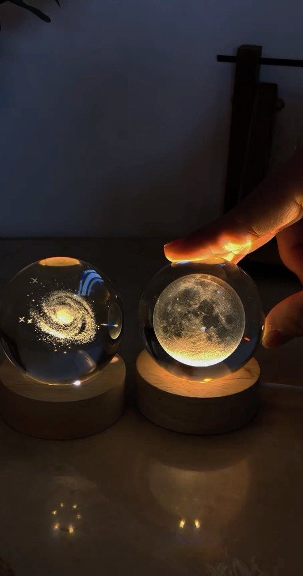 Two glowing crystal ball lamps with galaxy and moon designs on wooden bases.