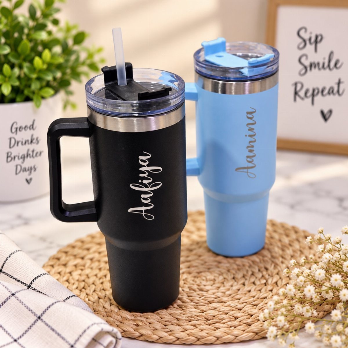 Two personalized insulated tumblers with names, one black and one blue, on a mat.