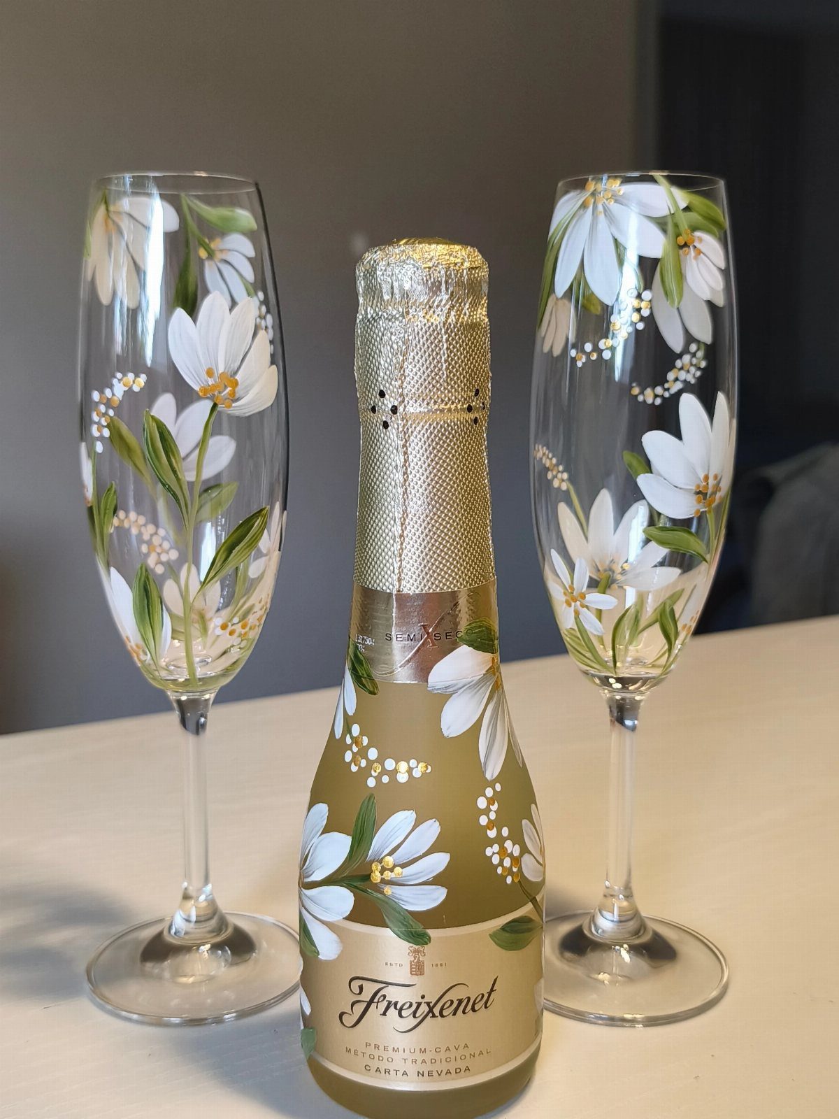 Hand-painted champagne flutes and a cava bottle featuring white floral designs and greenery.