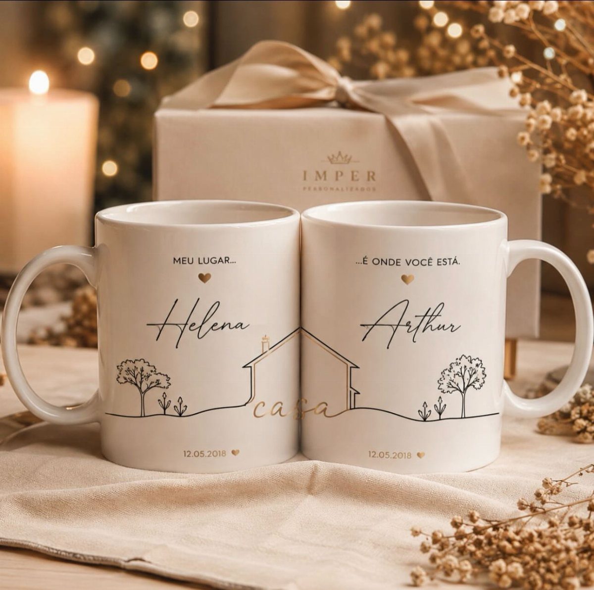 Two white personalized mugs with names and a house drawing on beige fabric.
