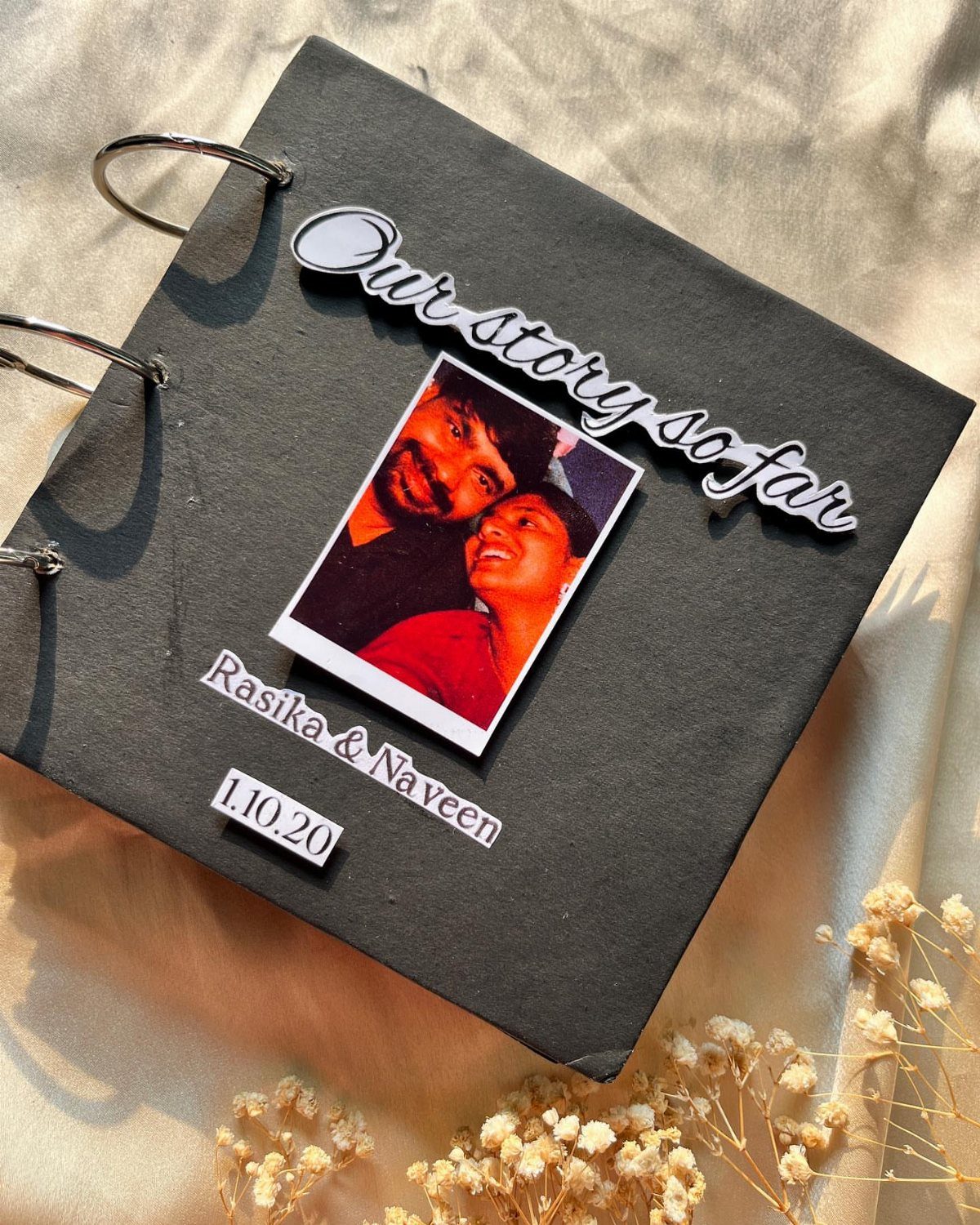 Black ring-bound scrapbook cover with a couple's photo and 'Our story so far' title.