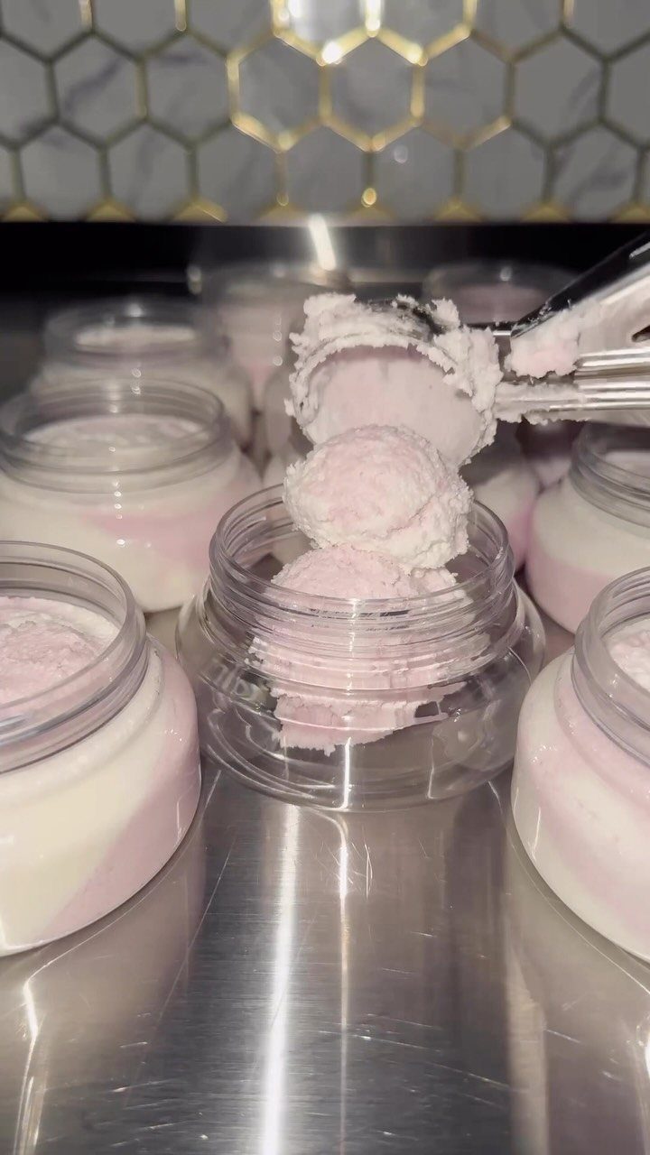 Whipped pink and white body butter in clear plastic jars on a metal surface.