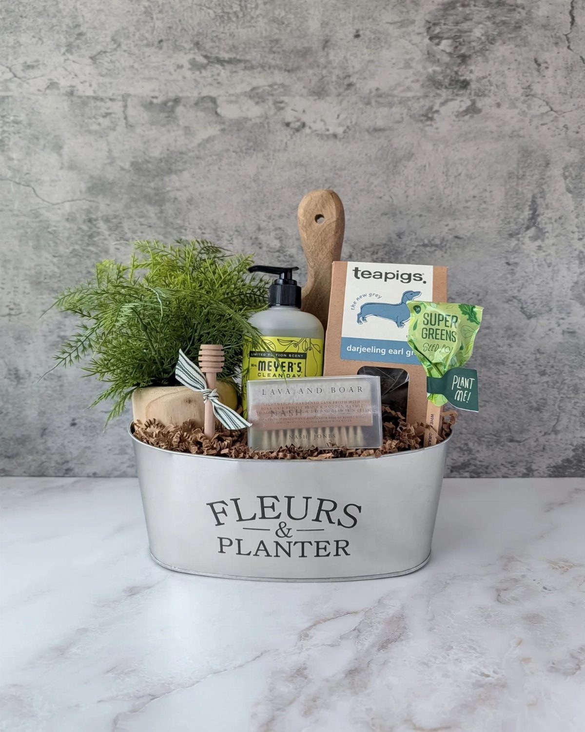 Silver metal bucket gift basket with a plant, soap, and tea on marble.
