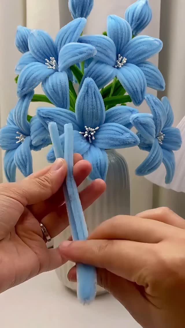 Hands crafting blue pipe cleaner flowers arranged in a grey vase.