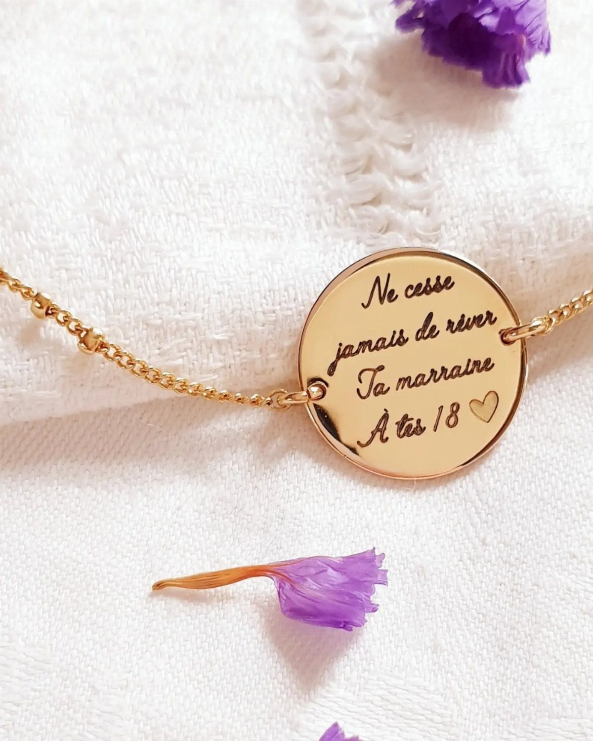 Gold circular pendant necklace with engraved French text on white textured fabric.