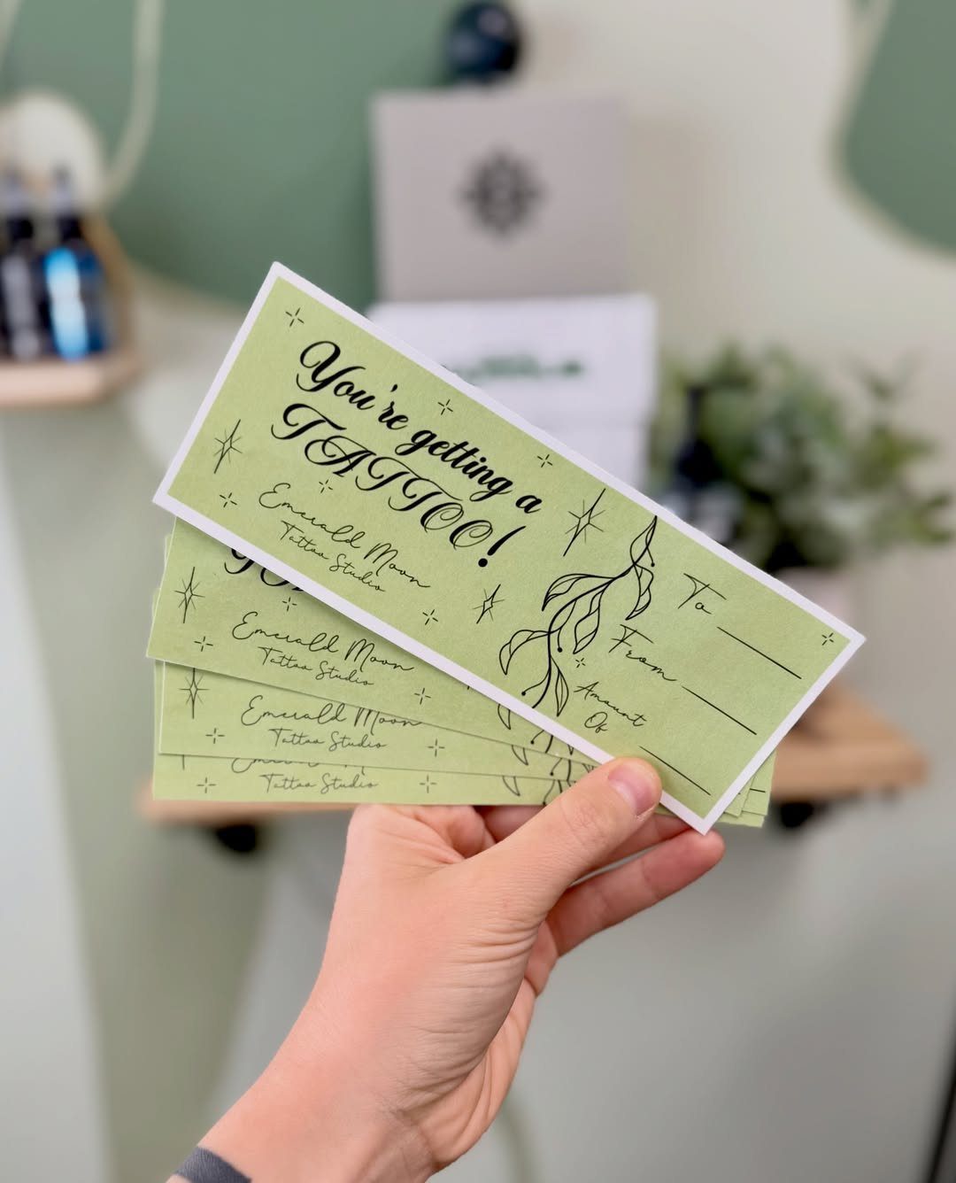 Hand holding a light green tattoo gift certificate with decorative text and illustrations.