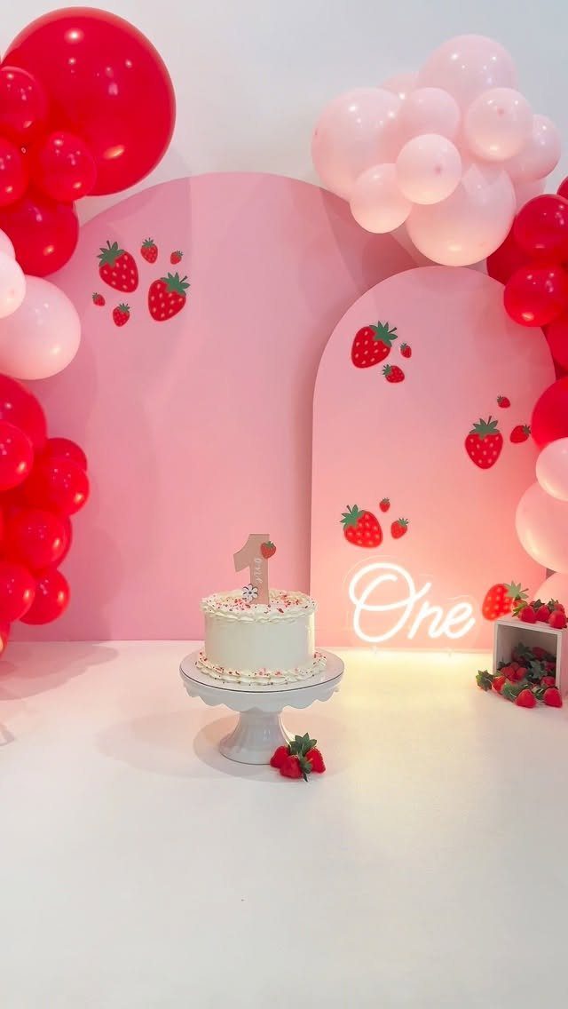 White birthday cake on a stand with strawberry decorations and pink balloons.
