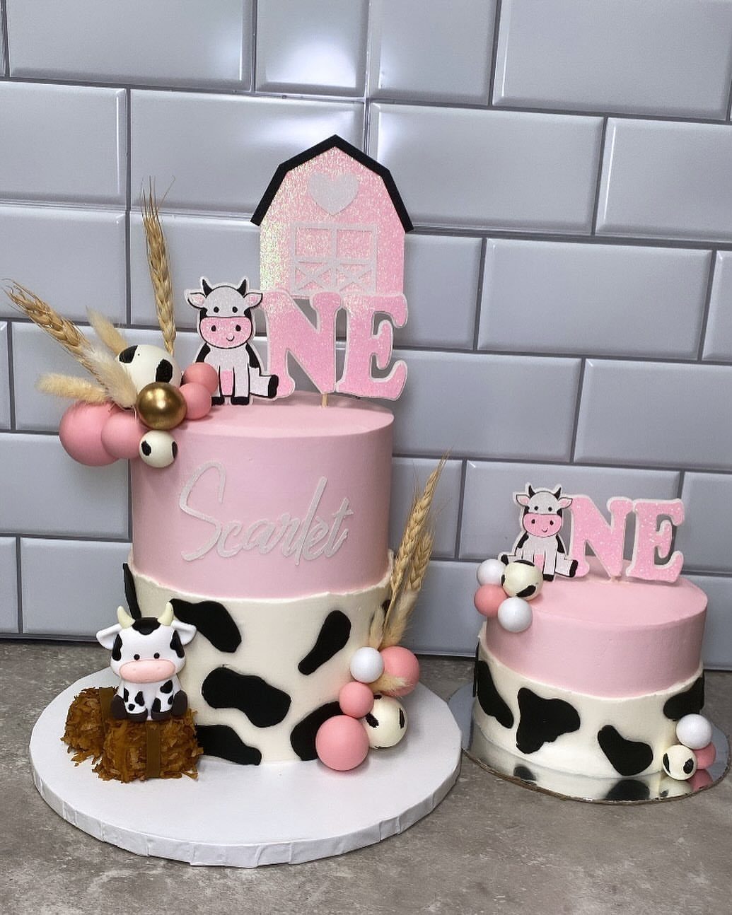 Two pink and white cow-themed cakes with barn toppers and cow figurines.