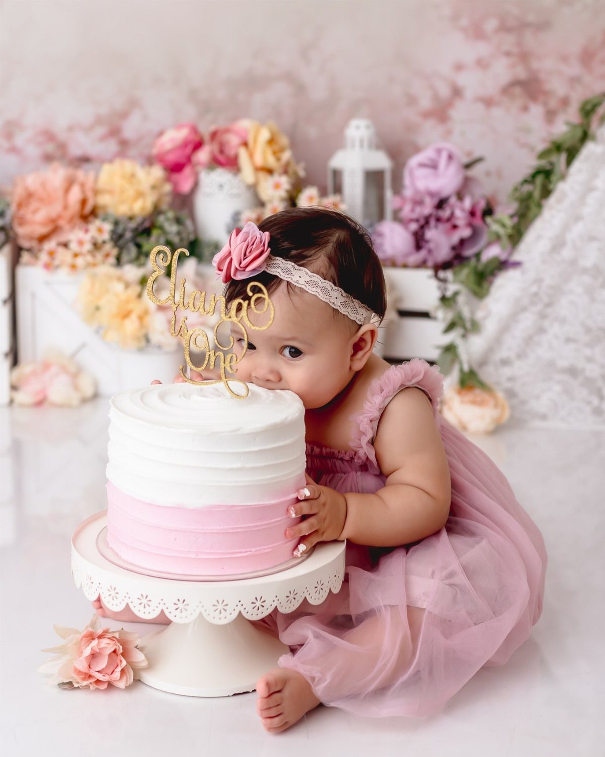Baby girl in pink tulle dress sitting by a floral cake smash setup.
