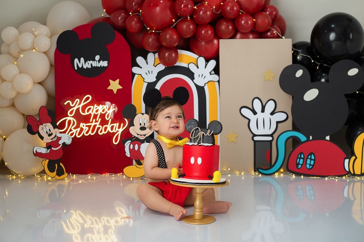 Baby in red outfit with a Mickey Mouse themed cake and decorations.