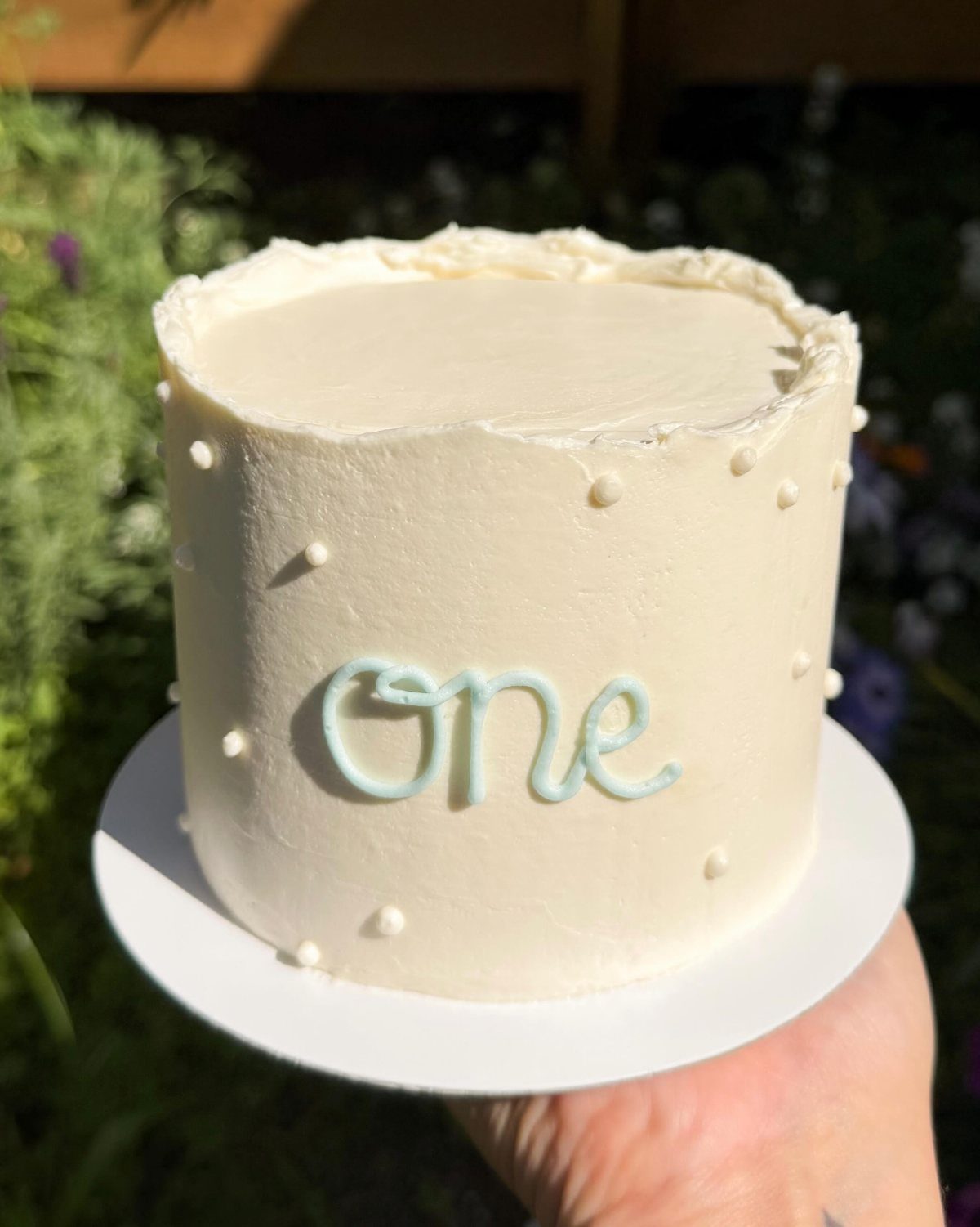 White frosted first birthday cake with light blue lettering and white dots.