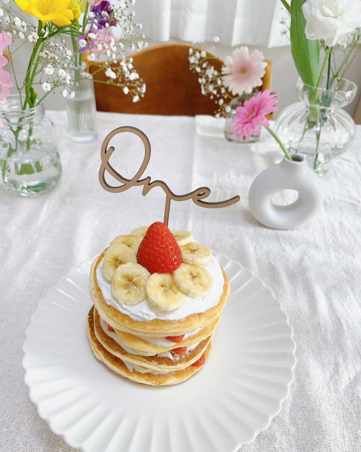 Pancake stack with cream, bananas, and strawberry topped with a wooden One topper.