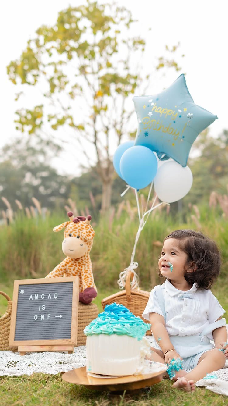 Baby boy with blue frosting on his face sitting by a cake outdoors.