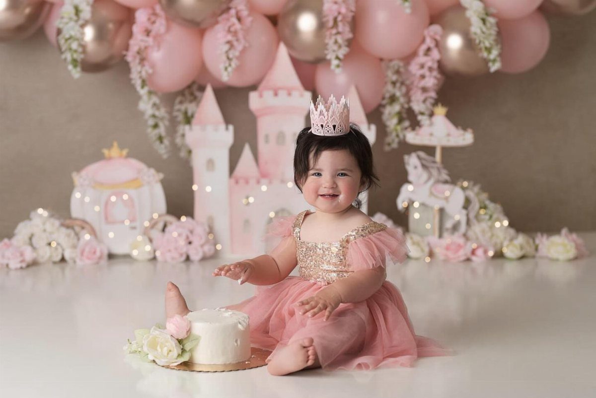 Baby girl in pink dress and crown sitting next to a white cake.