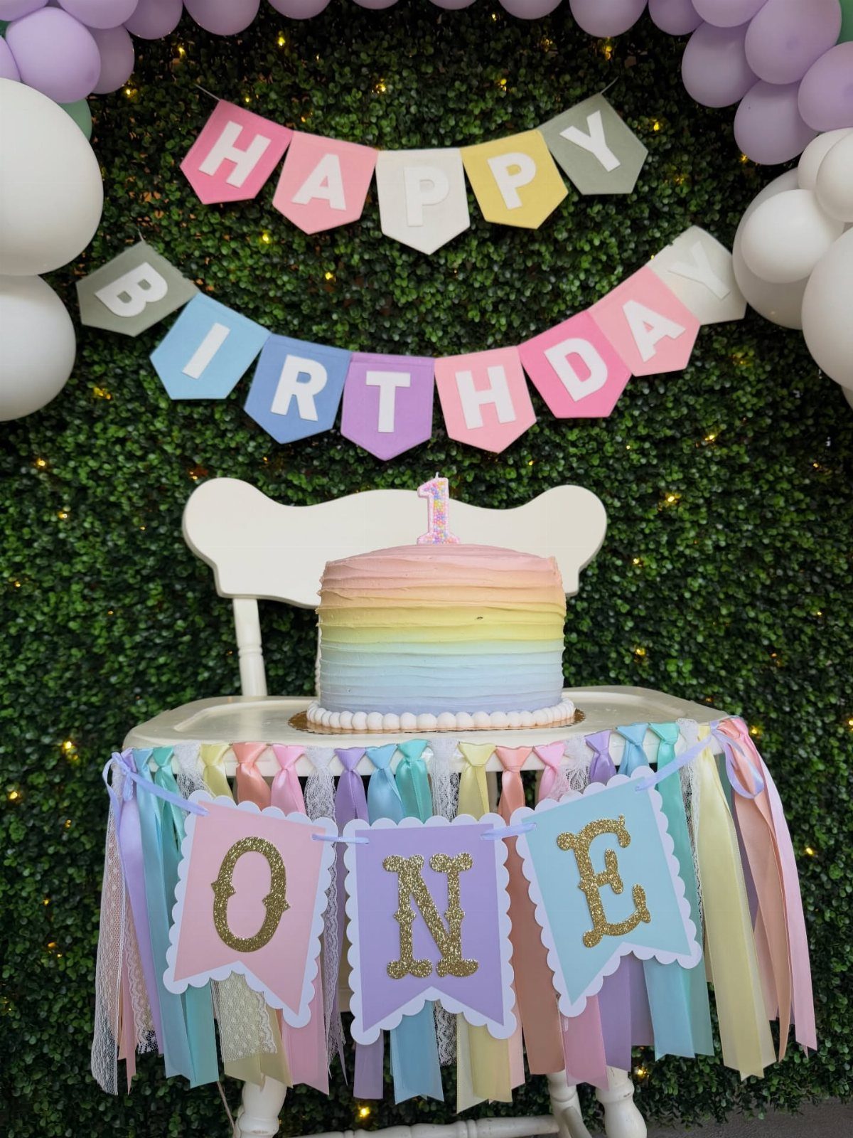 Pastel rainbow cake on a decorated high chair against a hedge wall background.