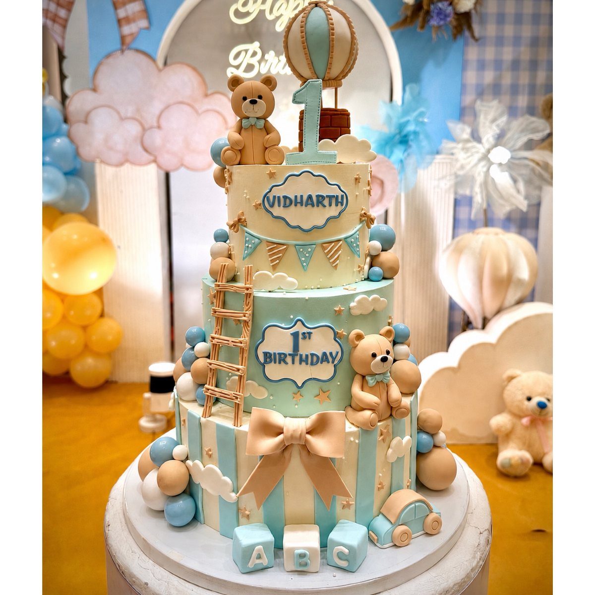 Three-tier blue birthday cake with teddy bears and matching cloud decorations in background