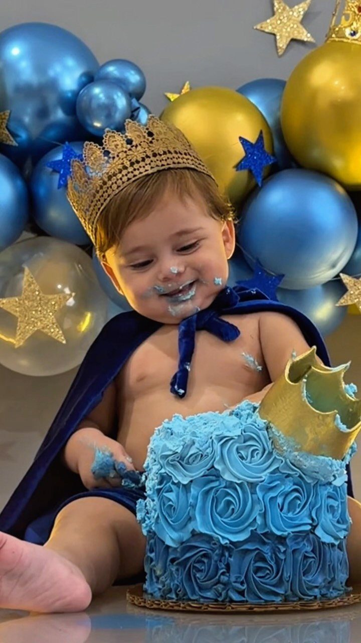 Baby in a crown and cape eating a blue rose-frosted smash cake.