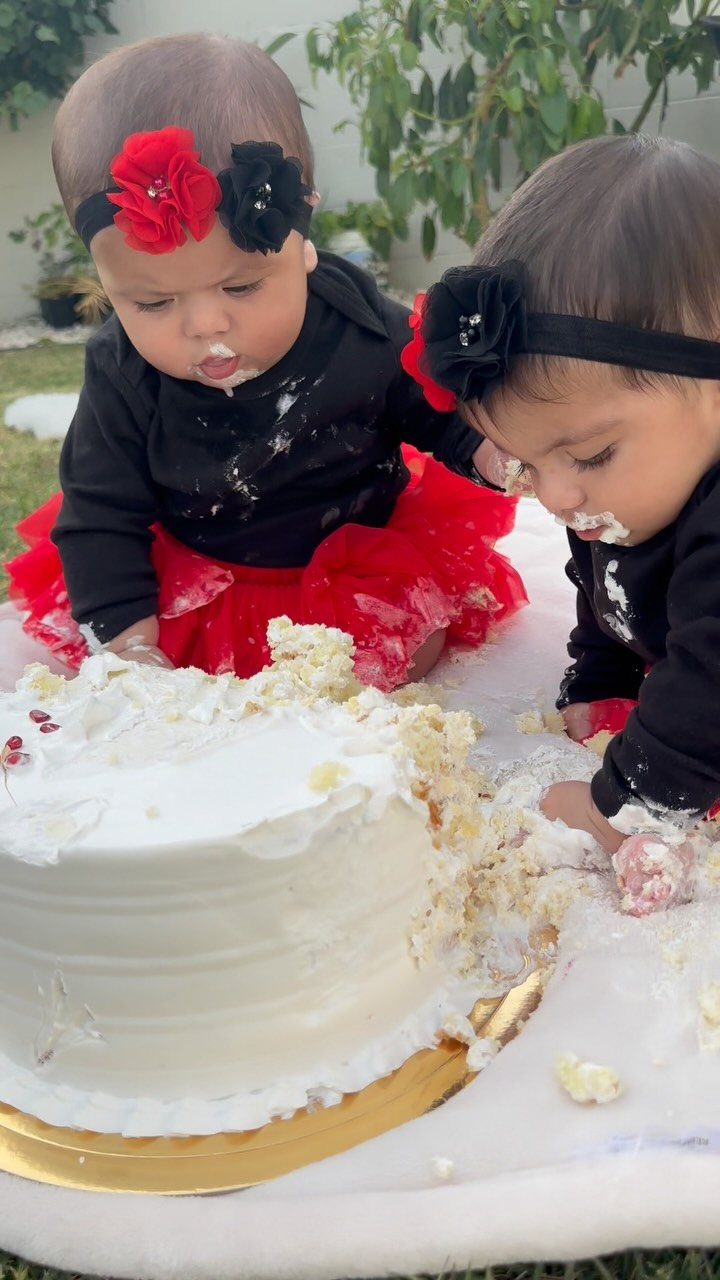 Two babies in red tutus smashing a white cake on a white blanket.