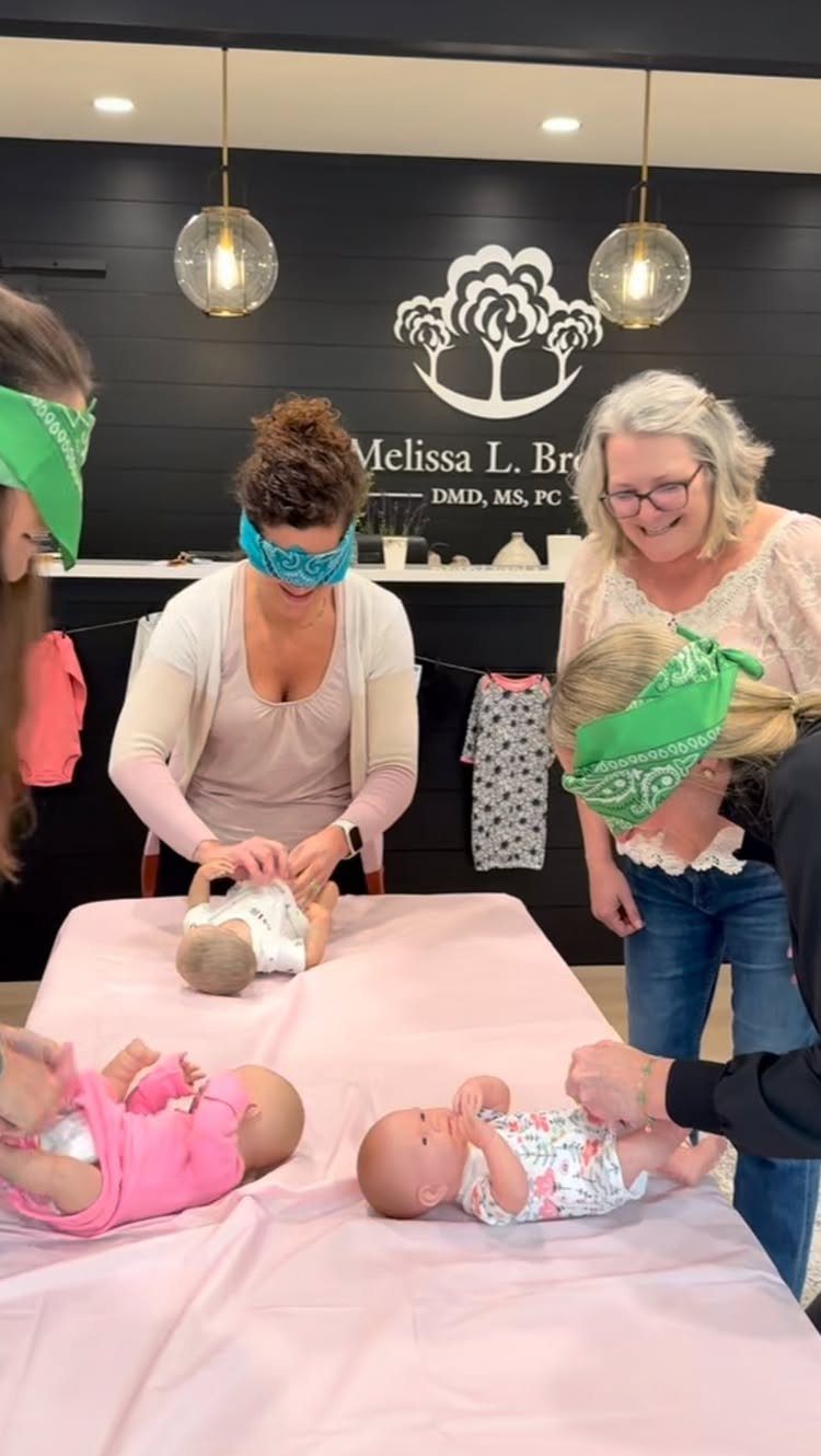 Blindfolded women and a girl changing diapers on baby dolls on a pink table