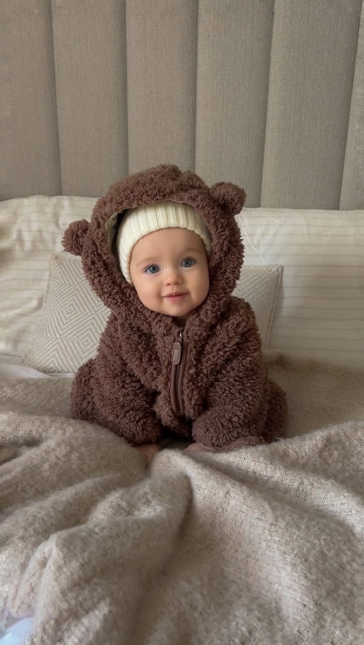 Baby wearing a brown fuzzy bear jumpsuit and cream beanie on a beige blanket