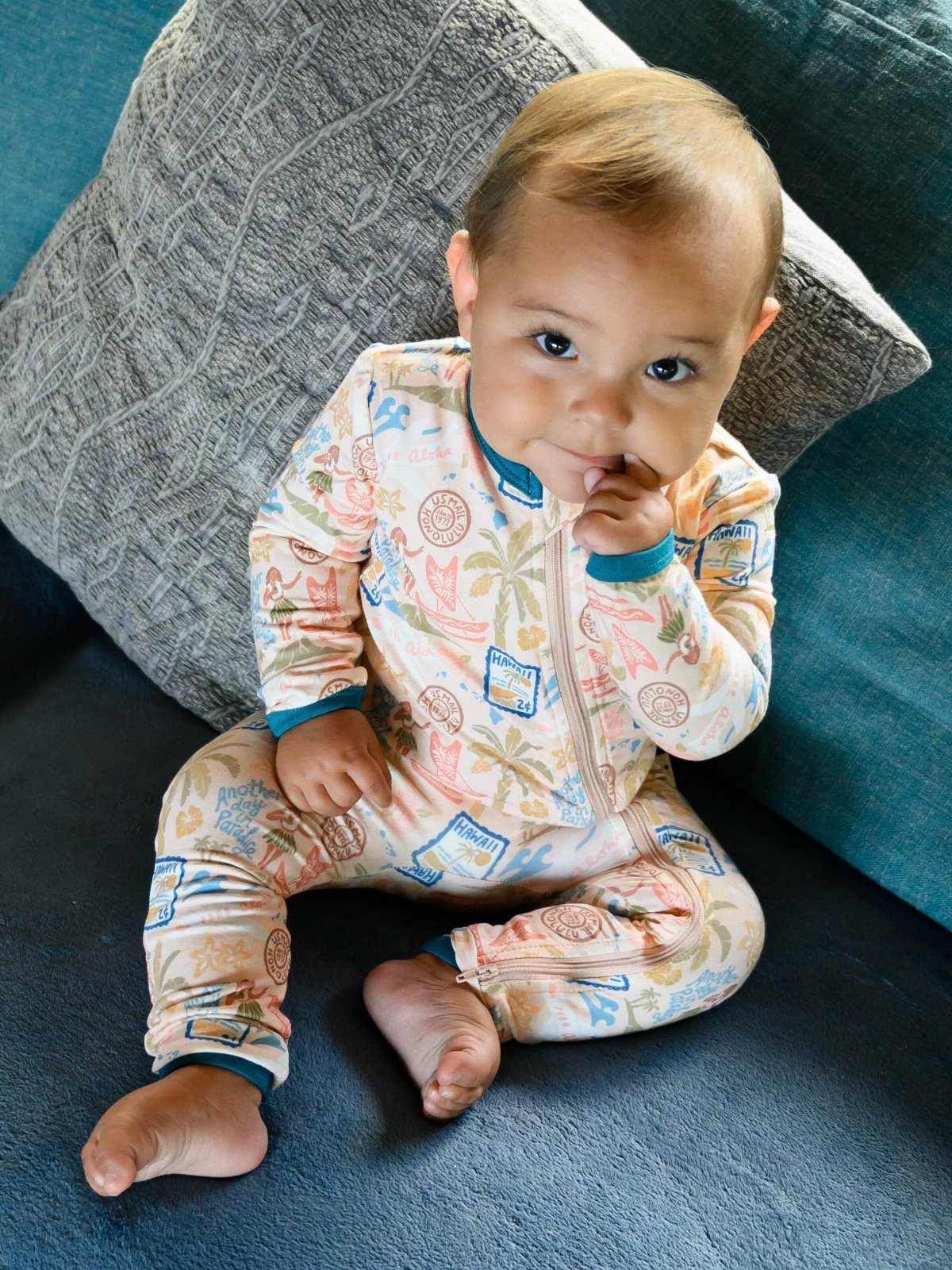 Baby sitting on a blue couch wearing a tropical print long-sleeved romper.