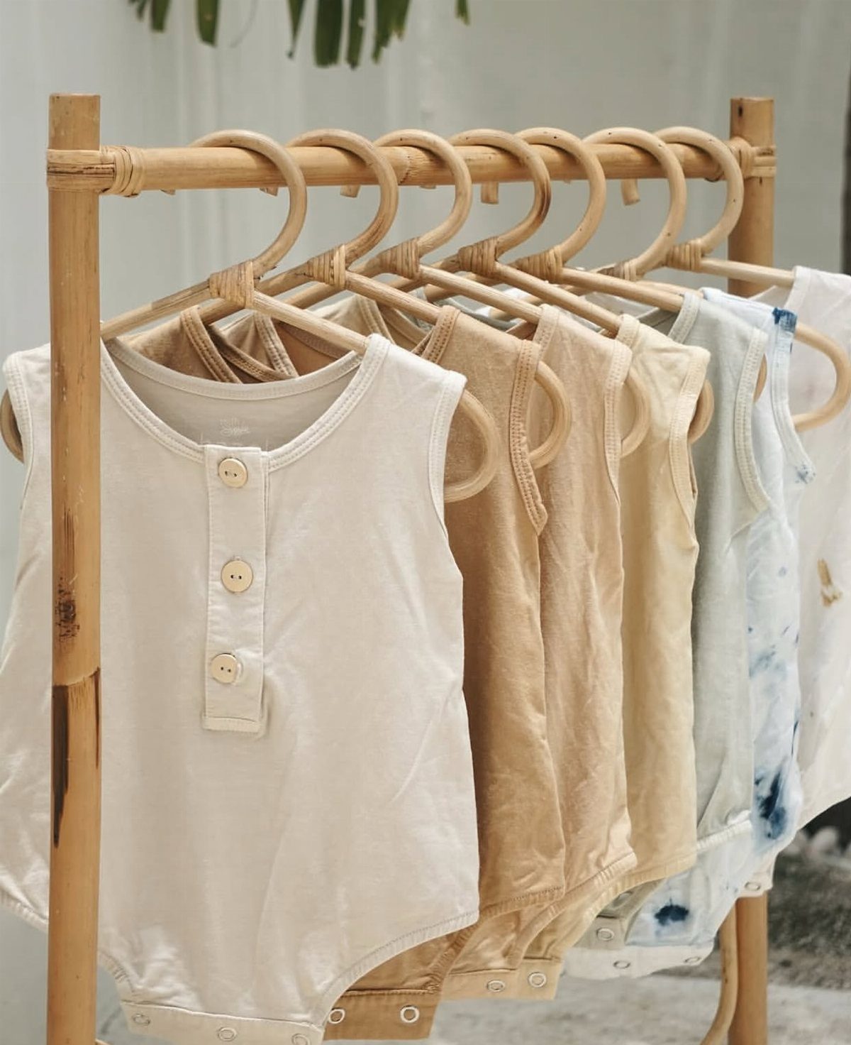 Sleeveless baby bodysuits in neutral colors hanging on a bamboo clothing rack.