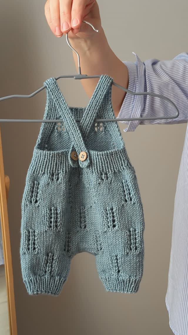 Light blue-grey knitted newborn overalls hanging on a grey hanger held by a hand.