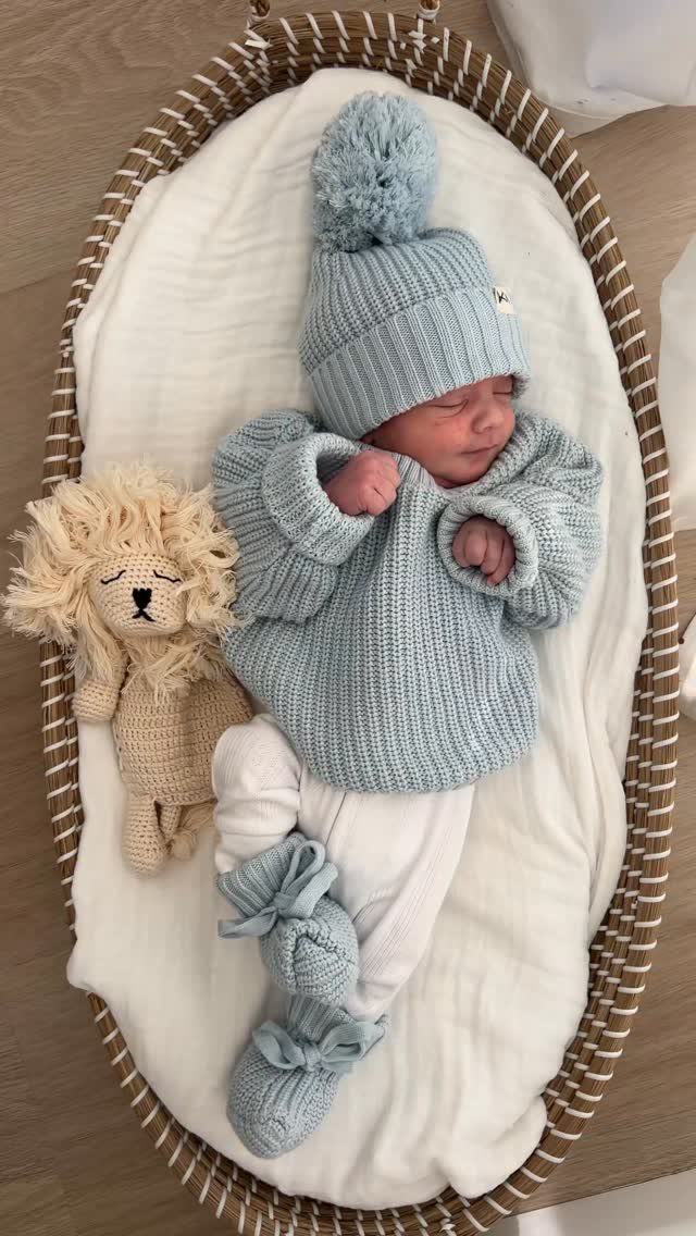 Newborn baby sleeping in a bassinet wearing a light blue knit sweater and hat.