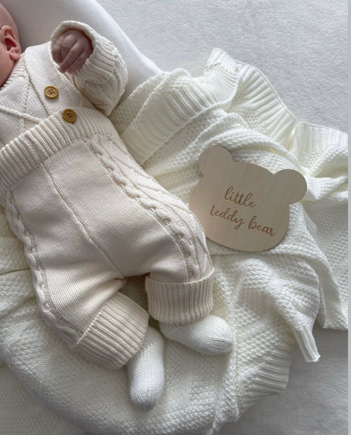 Newborn baby in a cream cable-knit outfit lying on a white blanket.
