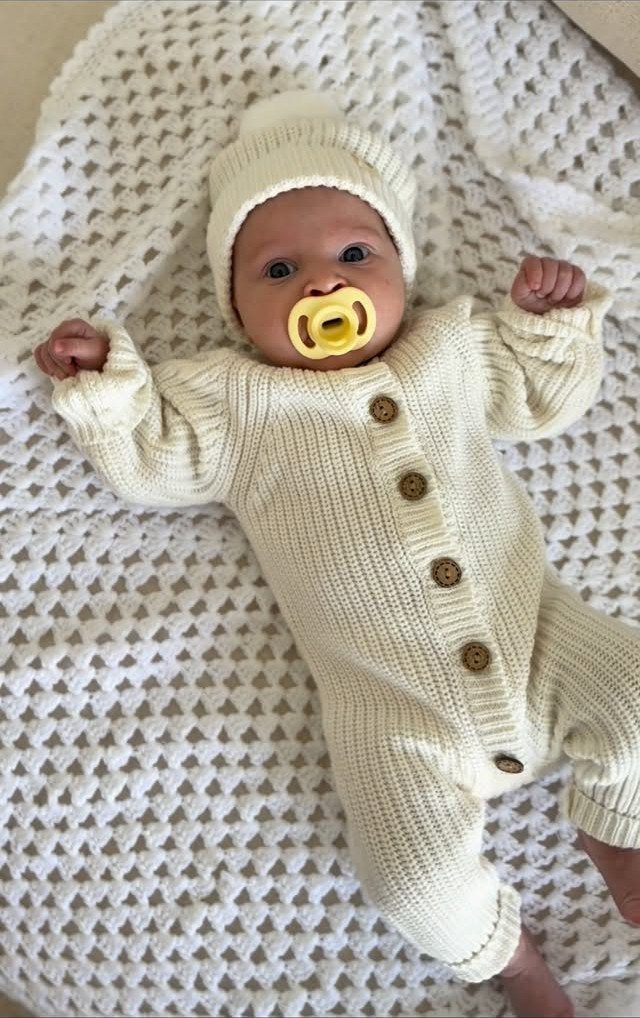 Newborn baby wearing a cream knitted jumpsuit and beanie on a white blanket