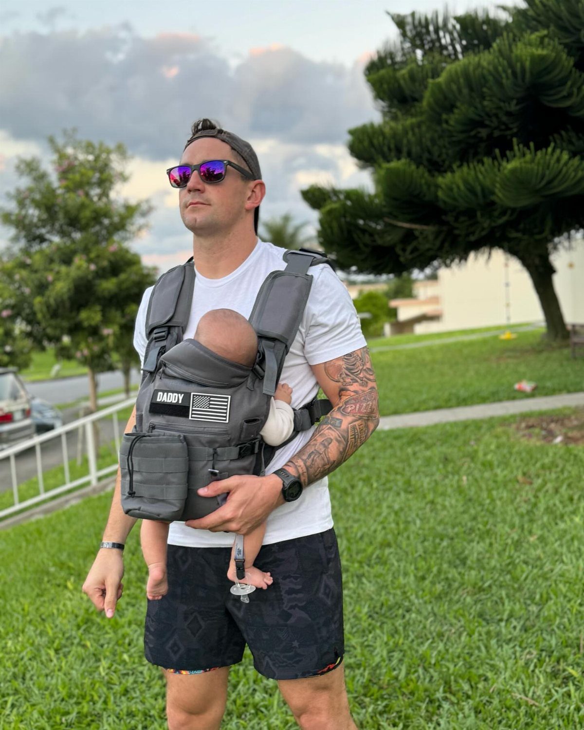 Man wearing a grey tactical baby carrier with a baby in a grassy field