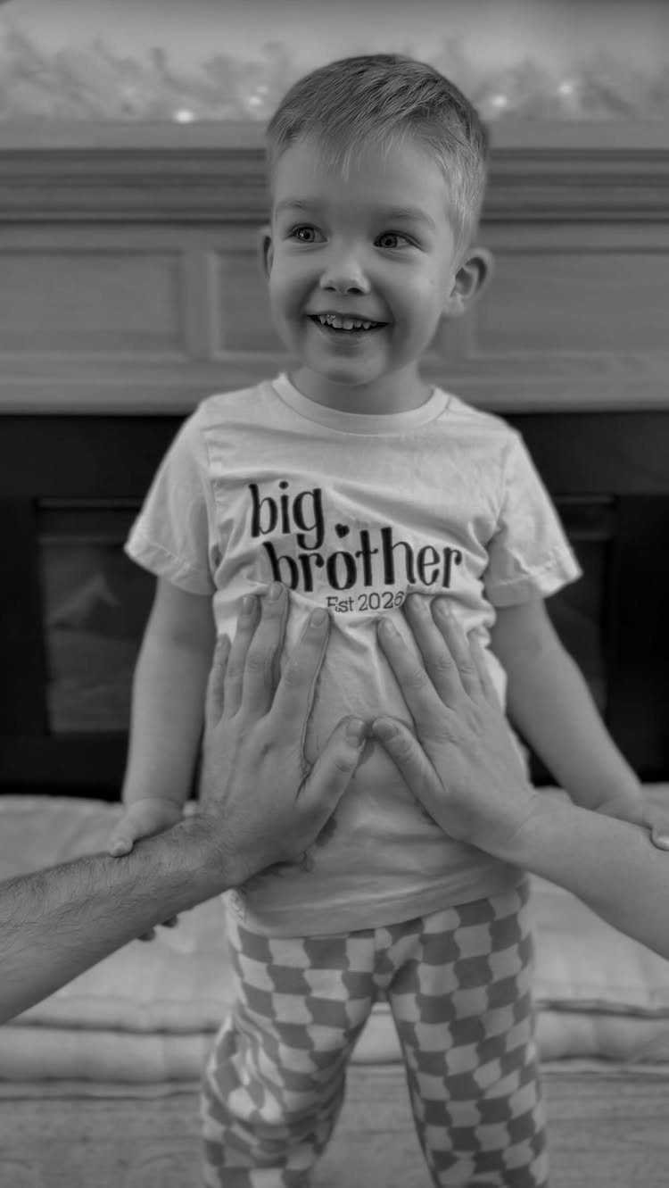 Smiling young boy wearing a big brother t-shirt with adults' hands on him.