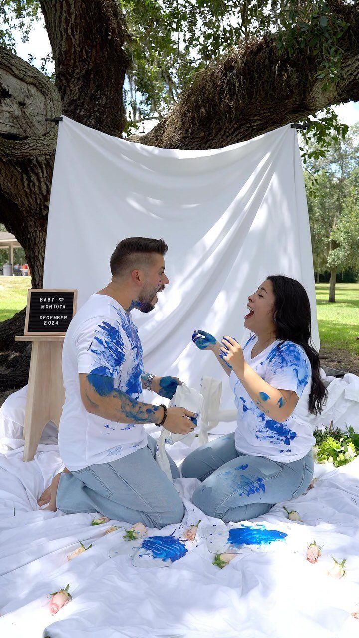 Couple covered in blue paint splashes laughing on a white sheet under a tree